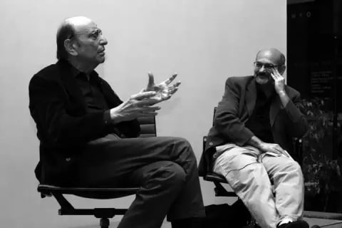A black-and-white photograph of two men who are seated and in conversation with each other.