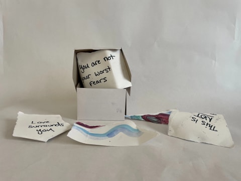 Depicts a white cardboard box with pieces of cardboard spilling out, phrases written on them. The community is invited to open the box of “Hope” and read through the phrases aloud.