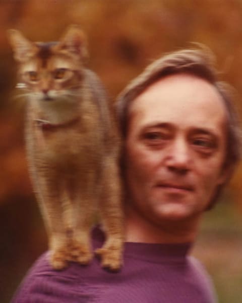 A photograph of a man in a purple shirt with a cat standing on his shoulder.