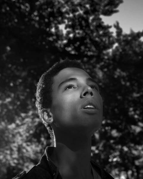 Black and White photograph of a young man taken from a slightly low angle against an out-of-focus background of trees. The man is looking up and his face is in shadow apart from a patch of light around one eye.  