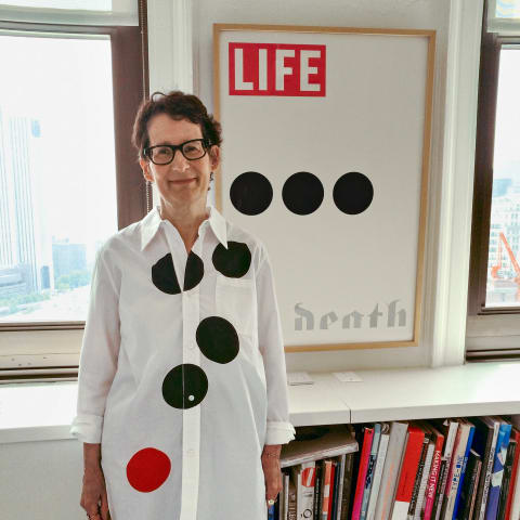 A photograph of a woman wearing a shirt with large polka dots and standing in front of a poster showing similarly-sized dots. 