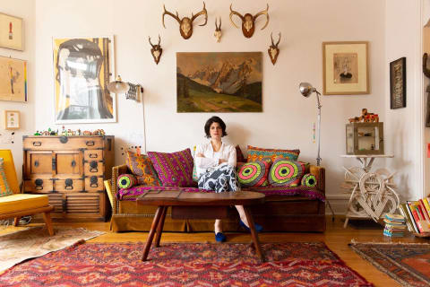 A portrait of a woman with dark hair sitting in a living room that is eclectically decorated with a variety of art and lamps and other curiosities..