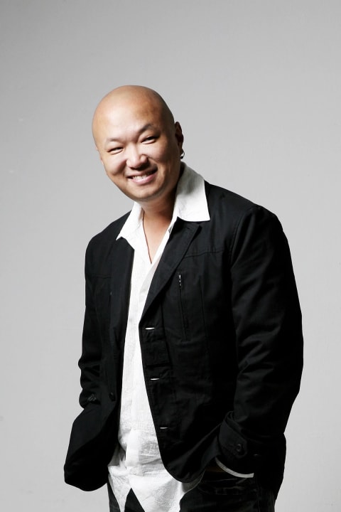 A bald, smiling Asian man in a black sports coat and white shirt standing in front of a gray background