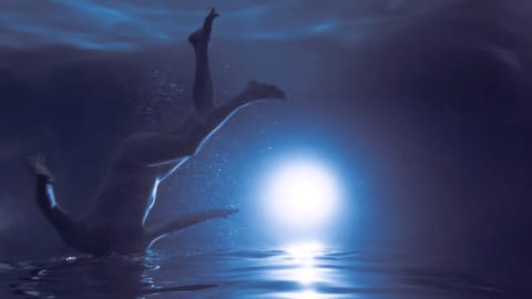 Photograph of a naked person swimming with their head out of the water, seen from below the water. The image is turned upside down and there is a bright light close to the water's surface that resembles a moon in the ocean horizon. The lighting makes the entire image look like its colored in blues and deep purple, with a white cool light in the middle. 