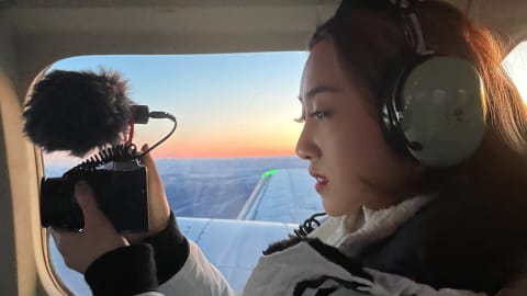 a woman sits in a plane, filming with a small camera. Out the window behind her, the sun sets over icy Alaska and the wing of the plane.