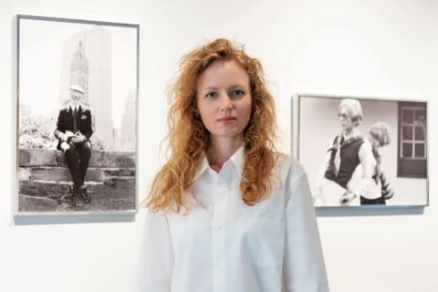 A woman with red hair stands in a gallery, behind her is a wall with two black and white photos on it 