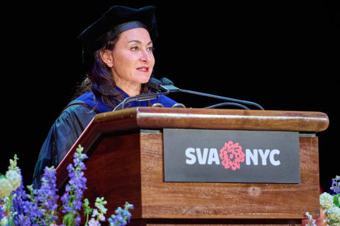 A woman wearing a graduation cap and gown at a dais, which reads "SVA NYC,"  and microphone, addressing an audience.