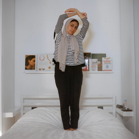 A woman stands on a white bed with her arms circled over her head. She is wearing a headscarf, striped shirt and black pants.