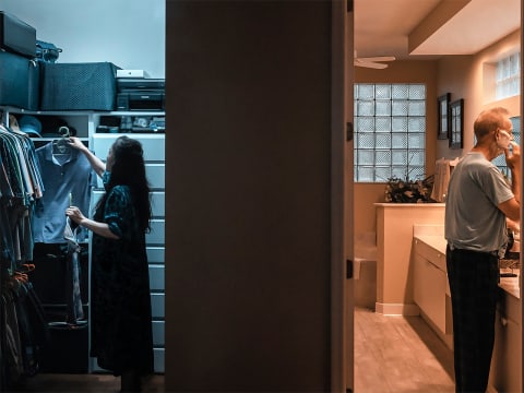 A dark wall in the middle of the image divides this color photograph into three equal parts. In the left third we see into a closet where an Asian woman is putting away a shirt in cool blue light. In the right third a white man is shaving in a bathroom lit by warm light. 