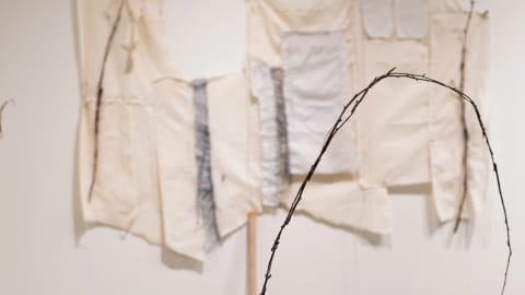 A formation of sticks tied together are mounted on a wooden block that sits on top of a pedestal. On the wall behind them hang beige and gray colored textiles that have been woven together.