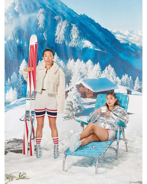 Two women are next to each other on a set designed to look like snowy mountains with a cabin in the back ground. One woman holds a pair of skis and the other reclines on a sun chair. 