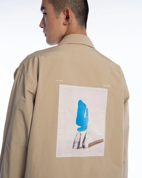 A man faces away from the camera wearing a beige jacket with a print of a painting on it
