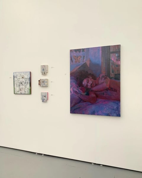 A photo of a white gallery wall with paintings hanging on it, one large purple one of a person reclining on a bed, and another set of smaller abstract ones
