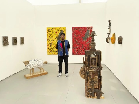 A young man stands in a white-wall gallery room filled with sculptures and paintings. Next to him is a taxidermy coyote with cotton buds covering it's body, behind him are two large paintings, one red and the other yellow 