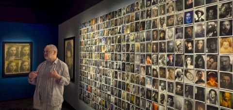 A man stands in front of a wall covered with photos in grid formation of portraits in various colors 