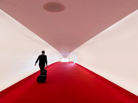 A man in a black suit walks down a long hallway towards an illuminated doorway. He is rolling a suitcase. The hallway is white with red carpet.