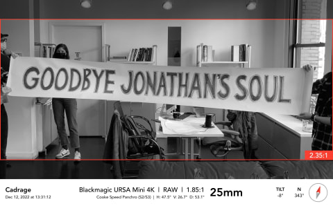 A screenshot of a still from a film featuring three people holding a sign that reads "Goodbye John's Soul" in an office space. It is in black and white.