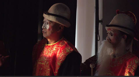 A still of two men waiting in the wings with beige hats, red vests, and white circles of face paint on 