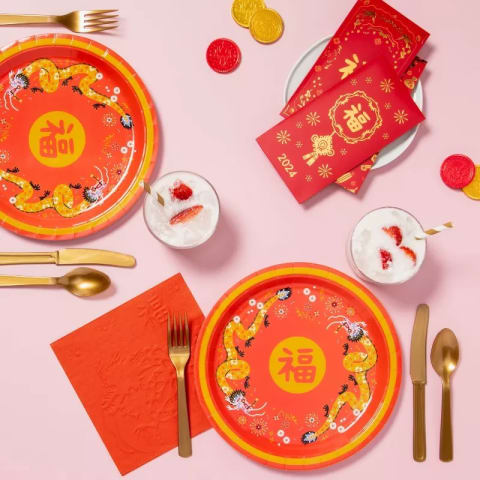 A pink background with paper and plastic cutlery laid out on it. The cutlery and plates are gold and red with dragon illustrations on them