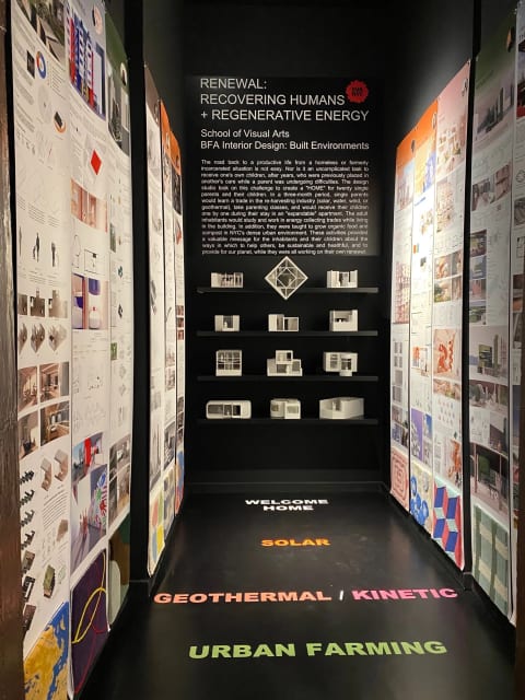 An installation in a narrow hall featuring images of interior design plans and patterns on the left and right walls. At the end of the hall are four shelves with white building models.