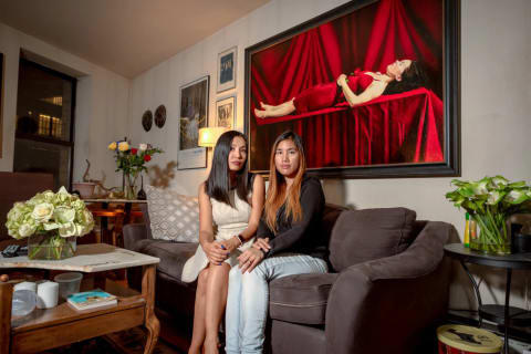 A photo of two women sitting on a couch in a living room. Behind them is a painting of a woman in a red dress laying straight on a table draped in red, with red curtains behind her. The living room is fully furnished.