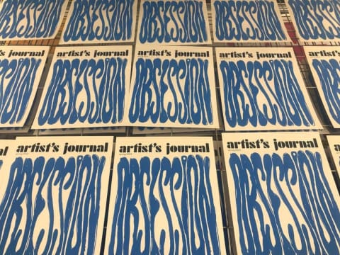 A photo of many magazine covers on drying racks with the word "obsession" printed on them in squiggled blue text  