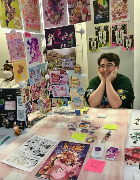 A person sits at a booth at a comics fair. There are comic books and stickers on the table in front of them and on stands next to them.