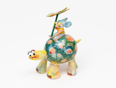 A colorful sculpture of a turtle with a little mouse figure riding on its back and holding a flower. The turtle is yellow and green and the mouse is blue and yellow, both with washes of other colors.