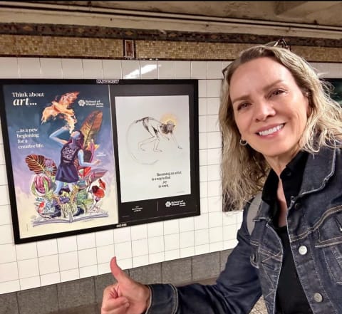 A woman with blonde hair poses in front of two subway posters. She is giving a thumbs up and smiling. Behind her, the poster on the left depicts a woman breathing fire into the shape of a bird, and to the right is a poster with a monkey on it.
