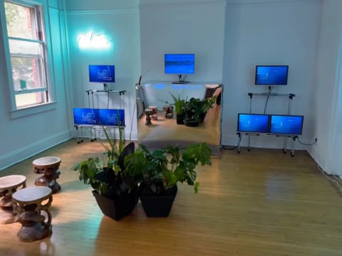 A dimly lit room containing 7 blue computer monitors and a neon sign that reads "water". There are potted plants in the center and three organically shaped wooden stools.
