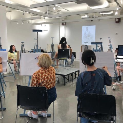 A group of students in a figuring drawing class draw a person sitting in a chair on a stage in the center of the room