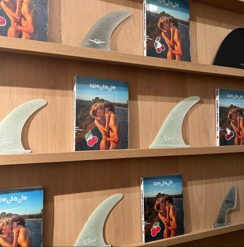 A wall of shelves with the same book repeated on it titled "spectacle." between each book is a flat fin-like sculpture.