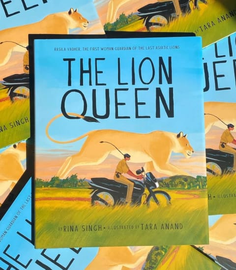 A book laid on top of a pile of several of other copies titled "The Lion Queen." The cover depicts a woman on a motorcycle and a large lionness behind her. 