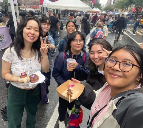 A group of young women at an open air market hold food and smile while taking a selfie