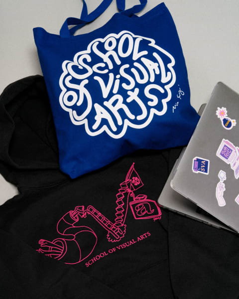 A blue tote bag that reads "School of Visual Arts" is laid out on top of a hoody with a pink illustration and a laptop with stickers on it