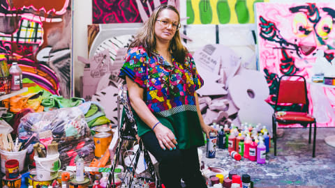 A woman leans against a stepladder with a can of spray paint in her hand, surrounded by colorful paintings and art supplies.