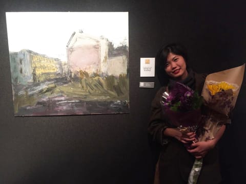 An artist posing with her art while holding two bouquets of flowers.