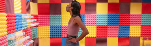 Profile view of a person wearing a pink crop top and a denim skirt with a black studded belt observes the room around them covered in textured tiles in colors of yellow, blue, teal, pink, brown & red
