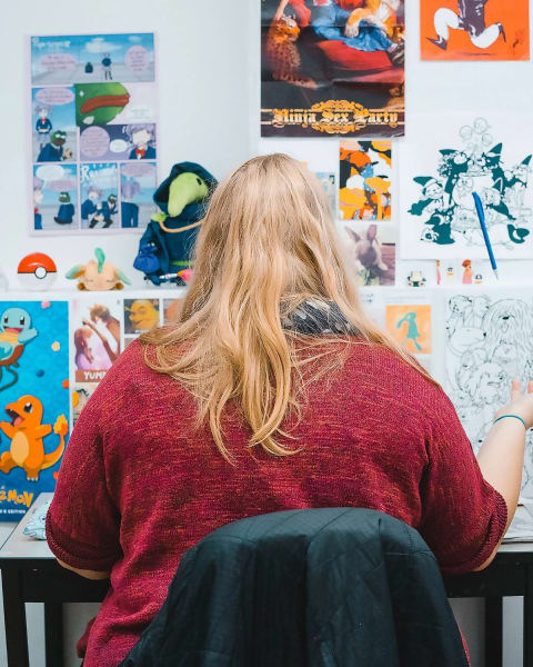 A person with long blonde hair, wearing a red sweater from behind. They're sitting in their studio surrounded by video game, Japanese animation and other kinds of paraphernalia. 