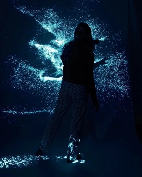 a person moves in front of a projection of abstracted blue particles in a dark studio space