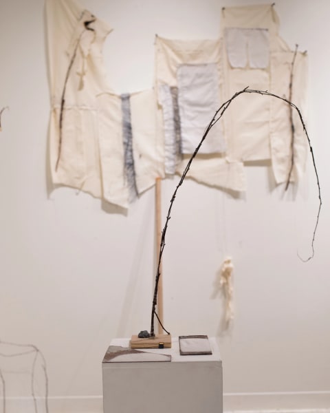 A formation of sticks tied together are mounted on a wooden block that sits on top of a pedestal. On the wall behind them hang beige and gray colored textiles that have been woven together.