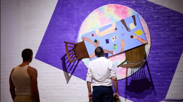 Two viewers inspect an art piece hanging from a painted brick wall.