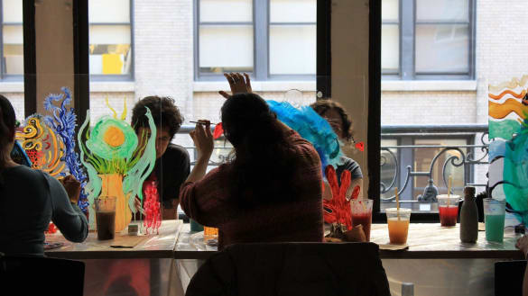 Artists paint on lucite panels while sitting down at a work table.