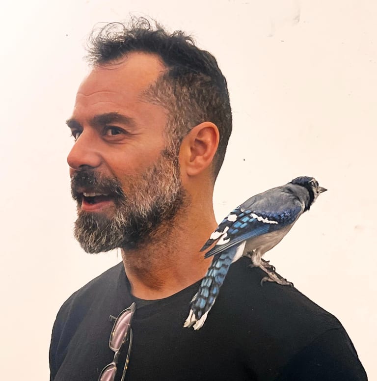 Man with blue bird on his shoulder standing in profile with a white background.