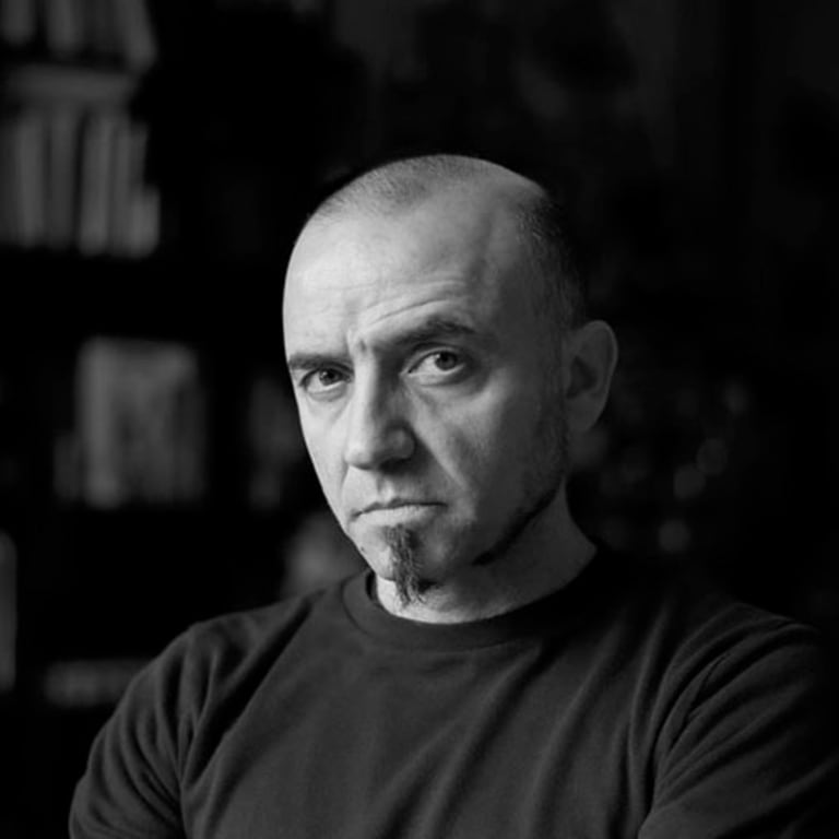 Faculty member Viktor Koen wearing a black shirt against a dark background and bookshelf.