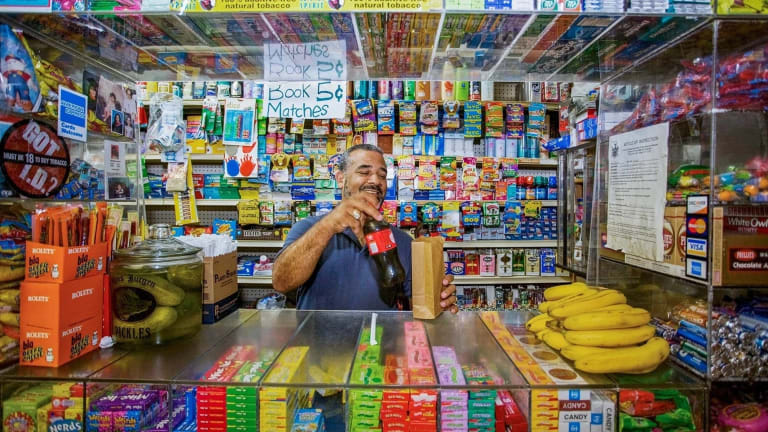 Portrait of a man behind a bodega counter. He is smiling and putting a soda into a paper bag.