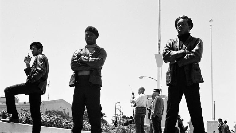 A B&W photograph of three members of the Black Panther Party in Black leather jackets Black berets standing guard outside with a flagpole in the background.