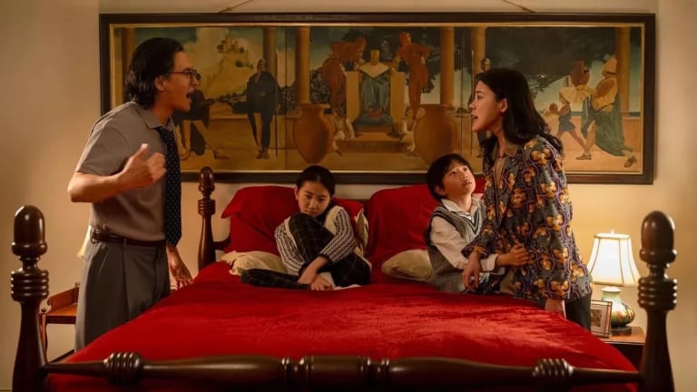 Photograph showing a large bed with a red bedspread; two children sit on the bed and their parents shout at each other from opposite sides of the bed.