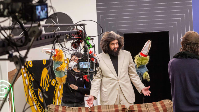 Production still from Jayson Musson's video performance entitled "His History of Art" from 2022. The artist is dressed in a beige suit with a black turtleneck and is performing as "Jay", and standing between two furry puppets, each wearing denim vests, sunglasses and pope's hats. There are production assistants and a camera on an arm in front of him, signaling that this is a behind the scenes shot.