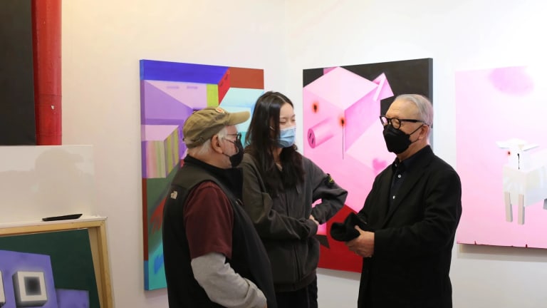 Three people wearing masks stand in a circle in front of multiple paintings 